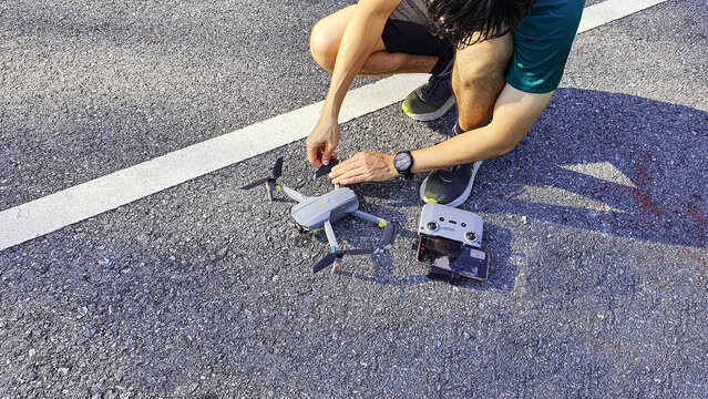 A person preparing and operating a compact camera drone on an asphalt road, with a remote controller and smartphone. Outdoor technology concept representing aerial photography