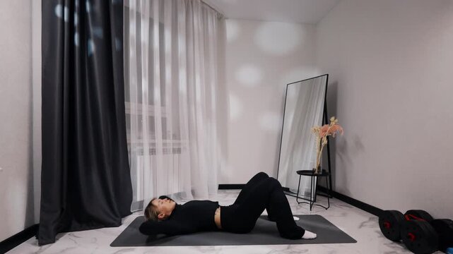 Young blonde woman in black activewear doing glute bridge during a home workout on a mat in a bright minimalist living room, side view