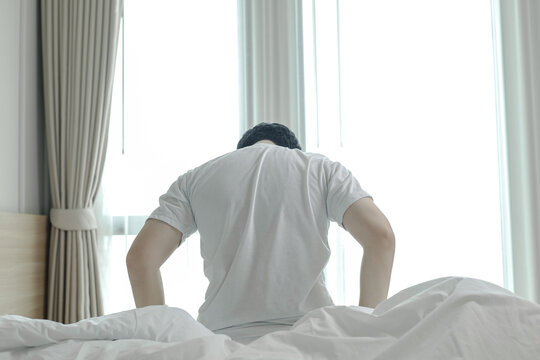 Rear view of a man sitting on bed in the morning, suffering from back pain, tiredness, or depression. Capturing health, fatigue, and mental wellness concepts.