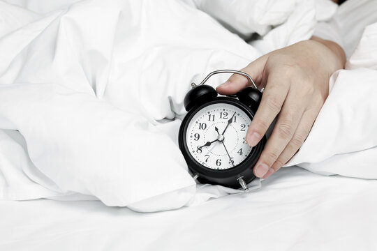 Close-up of hand reaching out to turn off or snooze black vintage alarm clock on white bed. Concept of morning wake up call, sleepy routine, time management, and late morning.