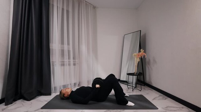 Young blonde woman in black activewear doing glute bridge during a home workout on a mat in a bright minimalist living room, side view