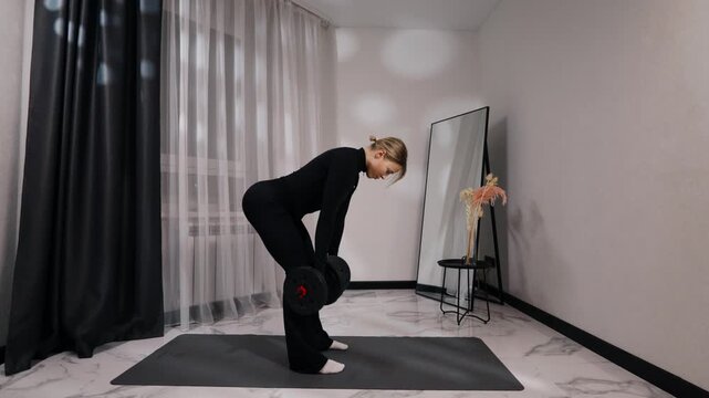 Young blonde woman in black activewear doing single-arm dumbbell row during a home workout on a mat in a bright minimalist living room, side view