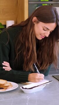 Tired young woman yawning and then writing down notes in her notebook while sitting at the kitchen table having breakfast, organizing her day and planning her schedule for better productivity