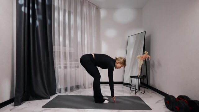 Young blonde woman in black activewear doing bodyweight forward bend during a home workout on a mat in a bright minimalist living room, side view