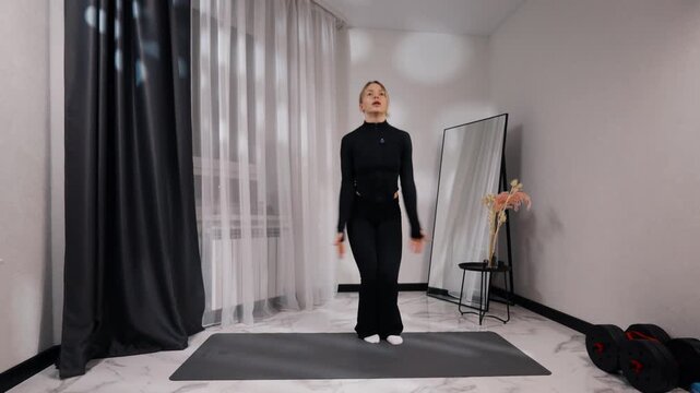 Young blonde woman in black activewear doing jumping jacks during a home workout on a mat in a bright minimalist living room, front view