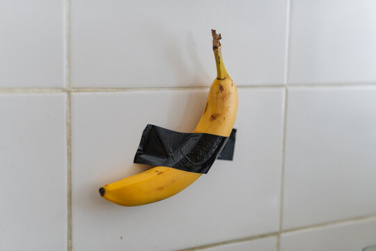 Banana with Black Tape on Wall. The image shows a single ripe banana affixed to a tiled wall with black tape around it. There is no additional context provided in terms