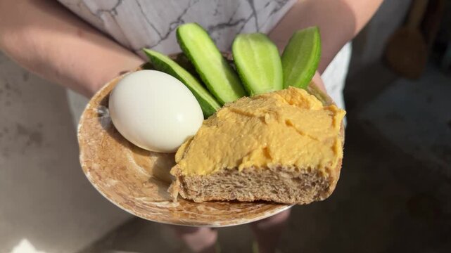 Person holding plate with boiled egg, fresh cucumber slices and bread with chicken or turkey spread, healthy breakfast snack, simple meal, nutritious food