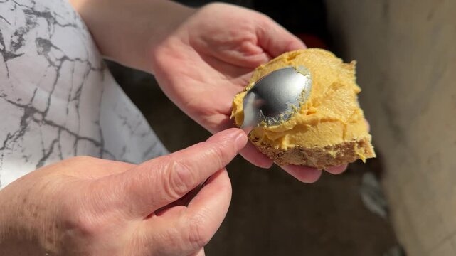 Hands spreading yellow peanut butter or cheese spread on bread with metal spoon, close-up of preparing snack, homemade food preparation, casual eating