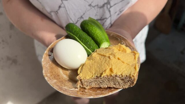 Person holding plate with boiled egg, fresh cucumbers and brown bread with meat and vegetable pate, healthy snack, simple nutritious meal, natural lighting