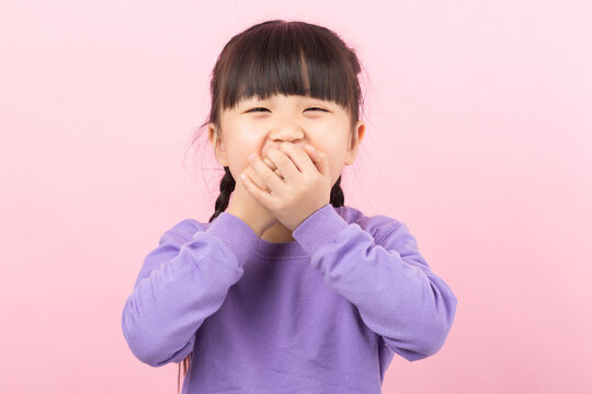 Cheerful little Asian girl covering her mouth with hands laughing on a pink background.
