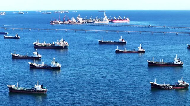 Fleet of oil tanker ships and commercial cargo vessels anchored in the deep blue sea near a long bridge. Global maritime logistics, international shipping trade, and energy transport.	