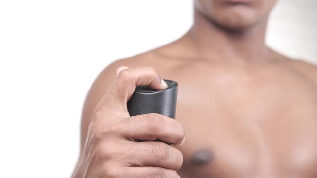 Young man using black deodorant spray can against a plain white background for body hygiene routine