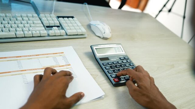 Person calculating budget with financial documents, Close-up of a person using a calculator beside printed financial papers, keyboard and mouse on a desk.