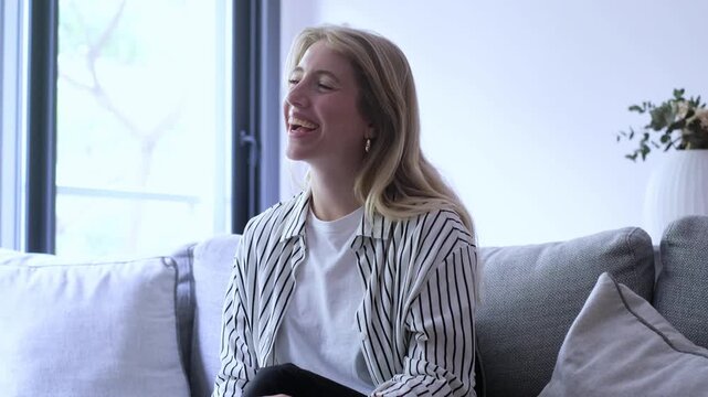Cheerful young blonde woman sitting on a comfortable gray sofa in a modern living room, laughing heartily during a pleasant conversation, expressing genuine happiness and positive emotions