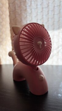 Pink portable usb fan spinning on a table with a whimsical cat design, providing a refreshing breeze and cooling air circulation for personal comfort on a hot summer day