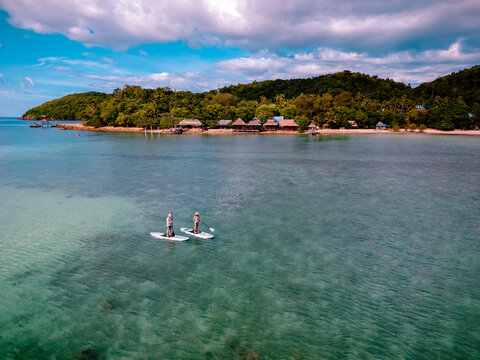 View of couple of tourist are enjoy on water sport paddle sup-board with kayaking along at the edge of the sea beach under crystal clear of water in the island of summer trip