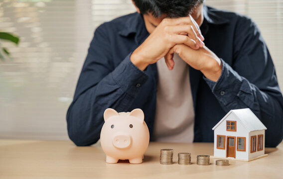 Stressed man holding his forehead, surrounded by a piggy bank, coin stacks, and a house model. Captures the struggles of money issues, financial risks, property concerns