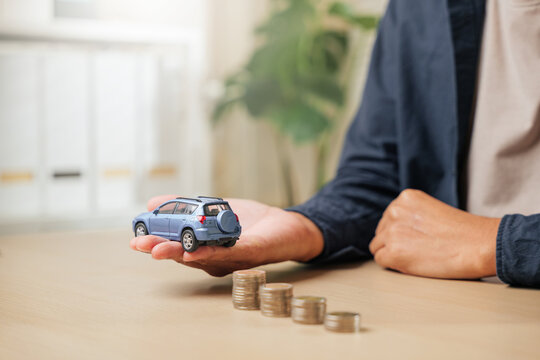 Close-up of a man hand holding a blue toy car model with ascending stacks of coins nearby. Highlights auto finance, car loan payments, credit cost, cash savings
