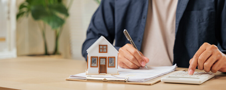 A man writes a signature on house insurance documents, using a calculator for tax and property details. Counting payments and rent by model house, he finalizes the price