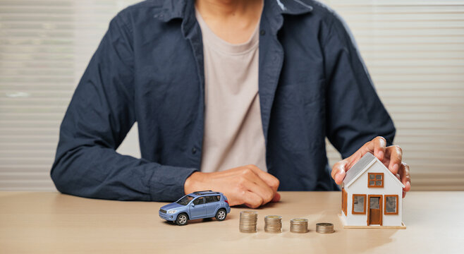 A person sits with hands clasped near a small model car, house, and coin stacks. Highlights finance, saving, property investments, auto and home loans, installment payments