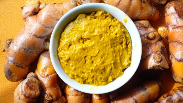 Fresh turmeric roots and bowl of golden turmeric paste on bright yellow background with slow motion camera movement