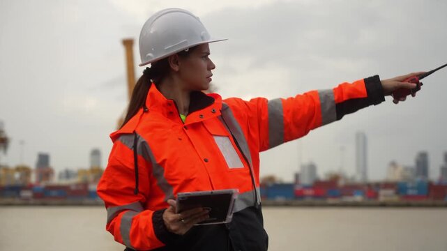 Female port engineer using walkie talkie and digital tablet managing operations at shipping container terminal, logistics manager in safety gear communicating via radio at harbor site