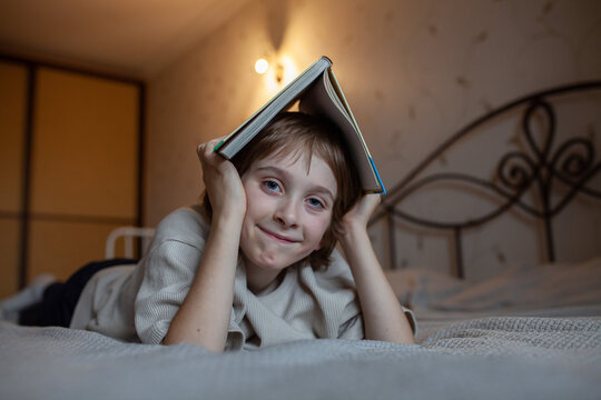 Cheerful boy lying on bed and holding open book over head like roof. Joy of reading, childhood creativity, cozy home life, curiosity, love for literature.