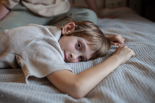 Gentle portrait of young boy lying on bed with soft natural lighting, looking towards camera.Childhood, coziness, relaxation, family lifestyle, home atmosphere