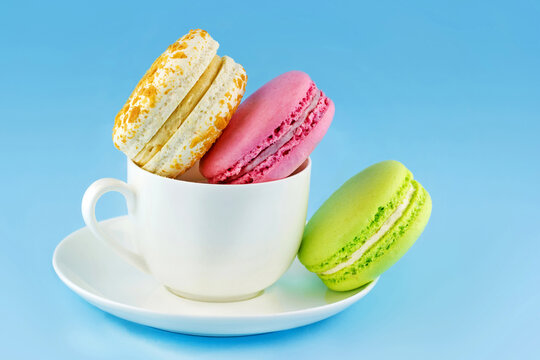 Three colorful macarons arranged in and beside a white coffee cup with saucer against a bright blue background, creating a cheerful dessert still life