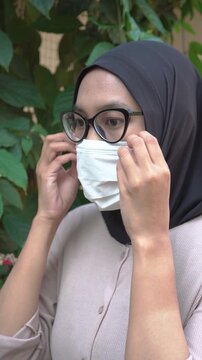 Side view of a modern Asian Muslim woman wearing a black hijab, cat eye glasses, and a protective white face mask outdoors.