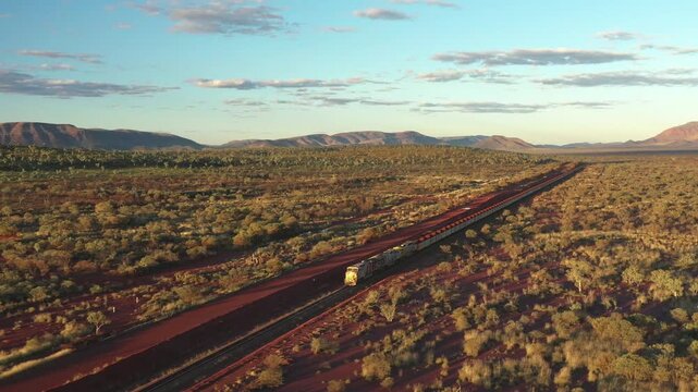 Aerial view of long freight train carrying iron ore on red dirt tracks through vast desert landscape under clear blue sky.