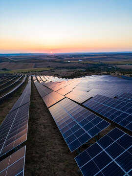 Solar panels in a large field capture sunlight at sunset on the horizon with rolling hills in the background