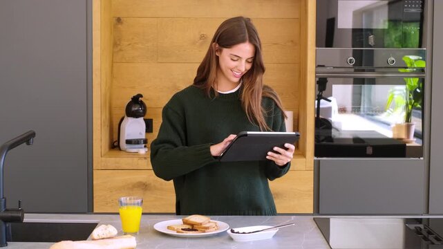 Beautiful young woman smiling while browsing on her digital tablet in a modern kitchen. She is standing by the counter, with breakfast items like toast and orange juice nearby