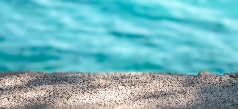 Close-up view of textured sandy surface with soft shadows against a blurred turquoise water background, creating a serene coastal atmosphere