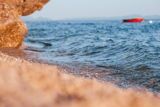Close-up view of sandy beach shoreline with gentle waves lapping against rocks and a small red boat anchored in the distance under clear blue sky