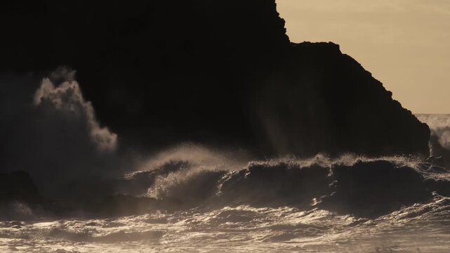 Dramatic ocean waves crash against a dark cliff face at sunset, creating a powerful and misty spectacle with golden light.