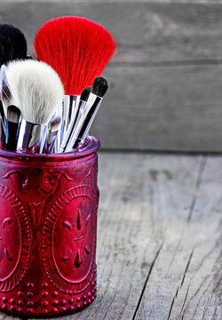 Closeup of assorted makeup brushes with soft bristles standing in a red embossed glass jar on a weathered wooden surface with neutral background