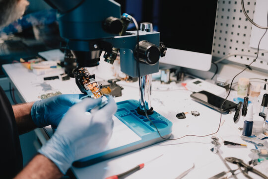 Technician examining and repairing miniature electronic components under a stereo microscope at a service bench, performing advanced diagnostics and precision micro repair