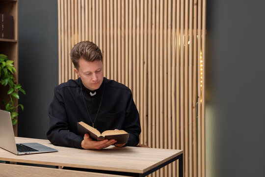 Priest in cassock reading Holy Bible at wooden table indoors, space for text