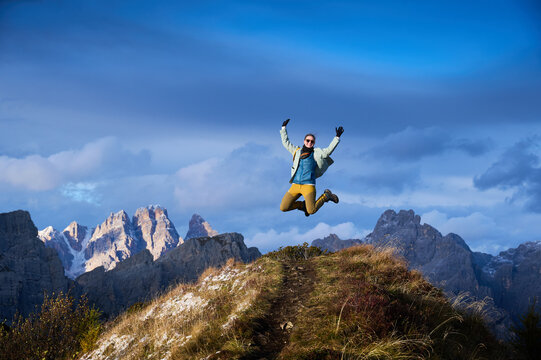 Joyful female hiker jumps with raised arms on snow-dusted alpine ridge in Dolomites, Italy, expressing freedom, achievement, positive energy, active travel and exhilarating mountain adventure