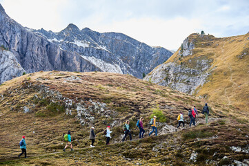Fototapeta premium Group of hikers climbs rocky alpine trail across open Dolomites terrain, surrounded by steep ridges, autumn grass, scattered snow. Conveys teamwork, endurance, active travel, guided outdoor adventure