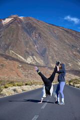 Fototapeta premium Female traveler helps friend hold playful handstand on mountain road near Mount Teide, expressing trust, teamwork and active friendship during Tenerife adventure in Canary Islands, Spain