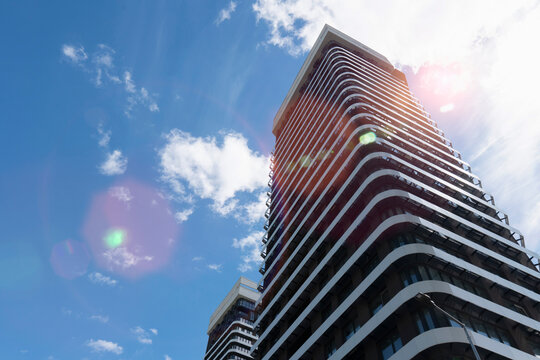 Low angle view of a modern high rise residential building with curved white balconies against a bright blue sky with white clouds and sunlight flare