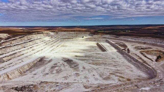 The scene shows a large open-pit mine with clear cuts in the earth. Heavy machinery is visible, and the sky is cloudy with some sunlight. The location seems to be in a rural area.