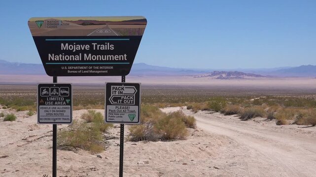 Mojave trails national monument sign at off-road trail entrance with vast desert landscape and mountains under clear blue sky.