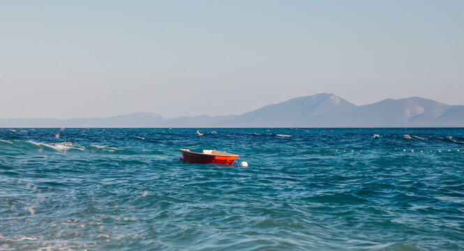 Small red boat floating on clear blue ocean water with distant mountains visible under a bright sky in a serene coastal landscape