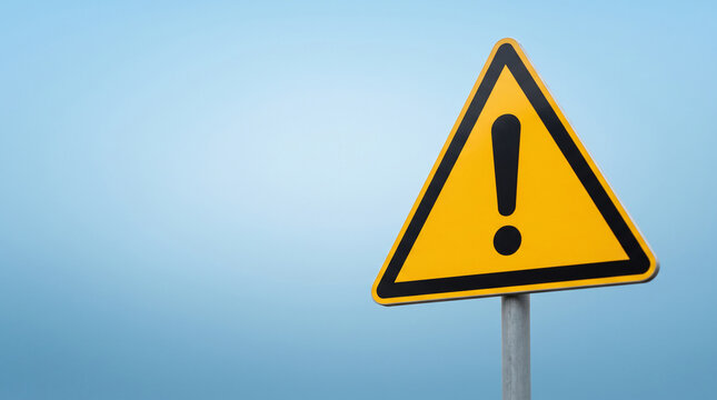 Yellow triangular warning sign with an exclamation mark, symbolizing danger or caution, against a light blue sky, offering free space for text.