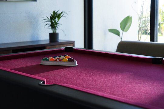 Magenta felt pool table holding metal triangle rack with colorful balls near windowed sofa