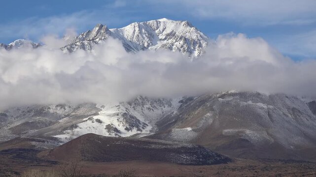 Snowy mountain peak partially obscured by moving clouds on a sunny day.