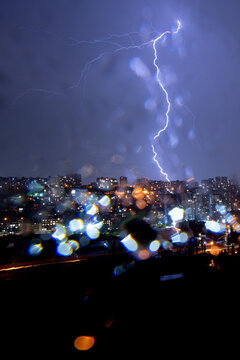 Dramatic vertical photo of a powerful lightning bolt flashing in the dark purple sky above a illuminated city skyline. Shot through a wet window with rain drops and bokeh highlights, capturing 